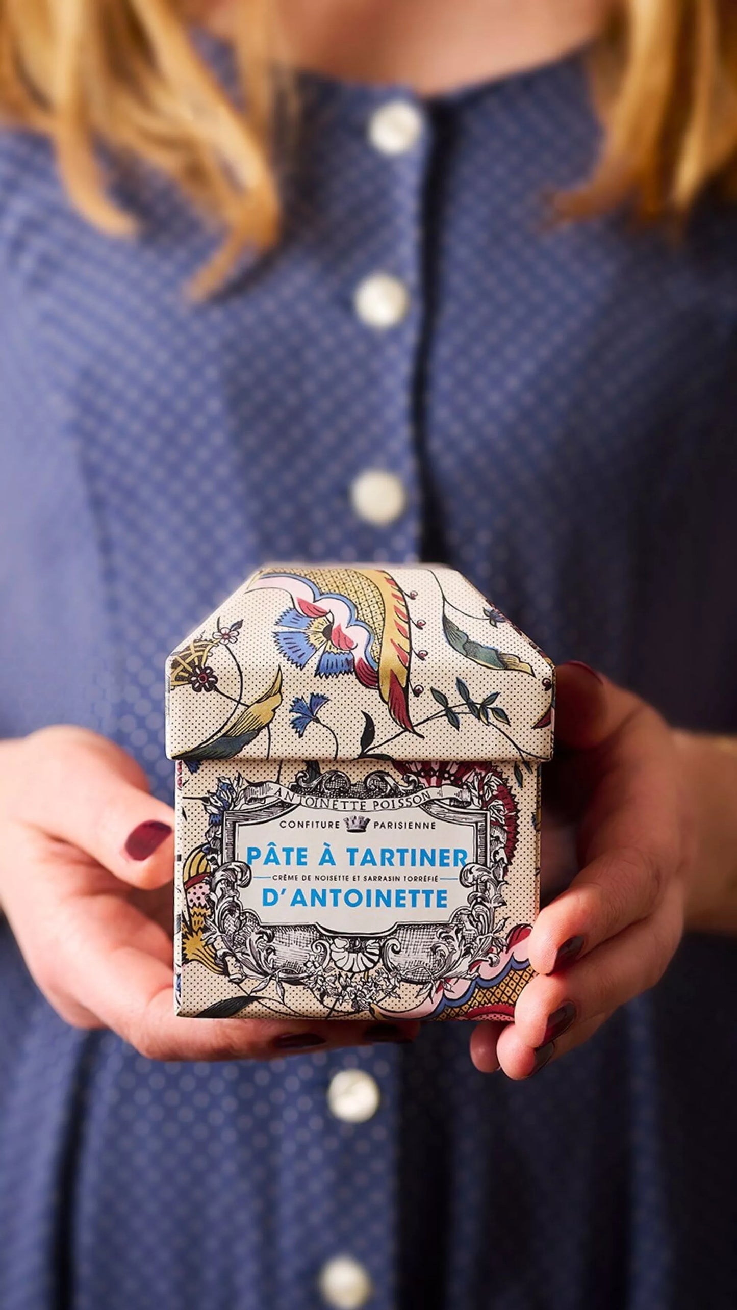 Person holding a decorative box labeled 'Pâçte à Tartiner d'Antoinette' against a blue background.