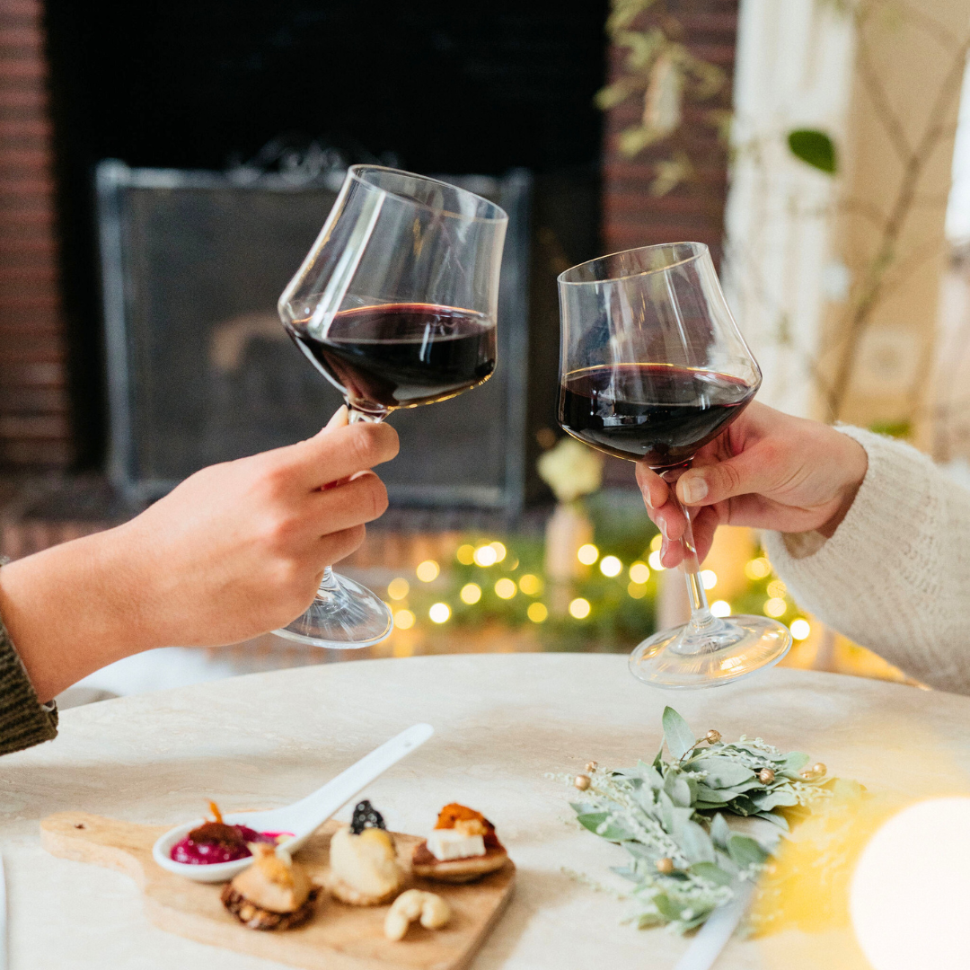 Two people toasting with red wine glasses in a cozy indoor setting with a fireplace and decorative lights.